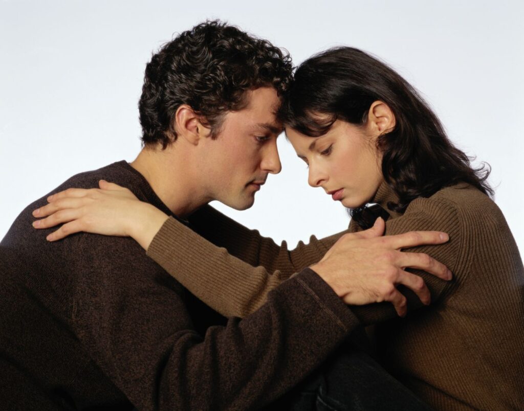 A couple, male and female, are leaning their foreheads against each other's while holding each other tenderly with somber looks on their faces. They are demonstrating being in each other's care while facing hardship, a stance that many couples facing trauma take.